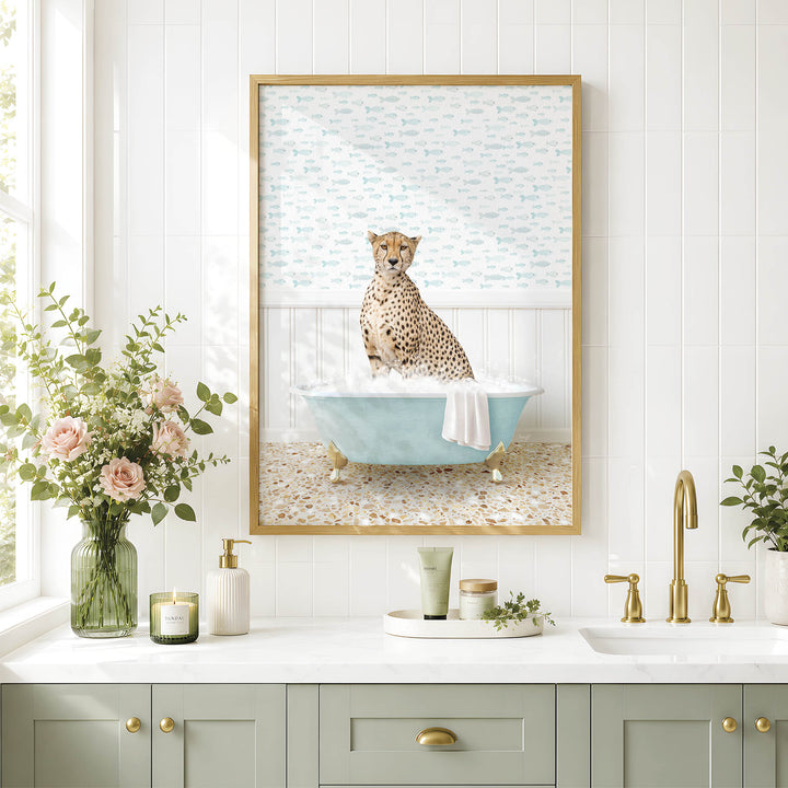 A framed image of a cheetah sitting in a bathtub, surrounded by flowers and candles, is displayed on a white tiled wall above a bathroom counter with a sink and gold faucets.