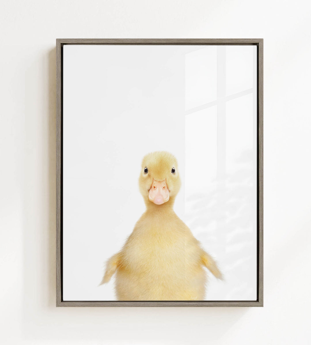 A cute yellow duck with a pink beak is the main subject of the framed image, which is hung on a white wall.