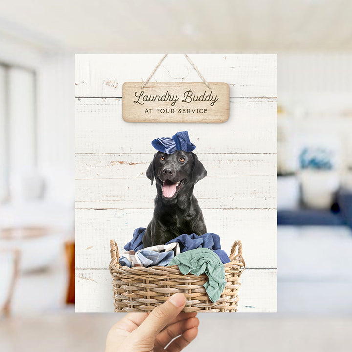 A black dog wearing a blue hat sits in a wicker basket filled with clothes, with a wooden sign above that reads "Laundry Buddy at your service".