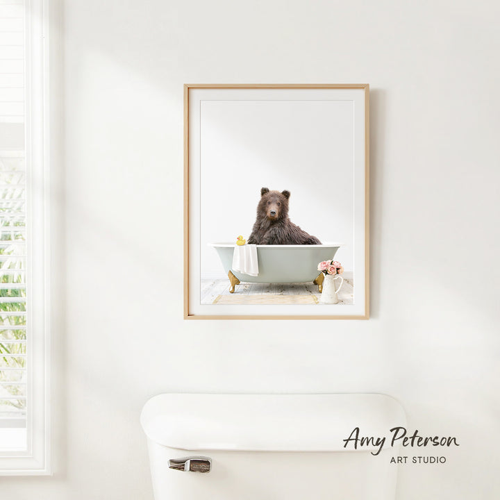 A framed image of a brown bear sitting in a bathtub, with a vase of flowers and a white toilet nearby.