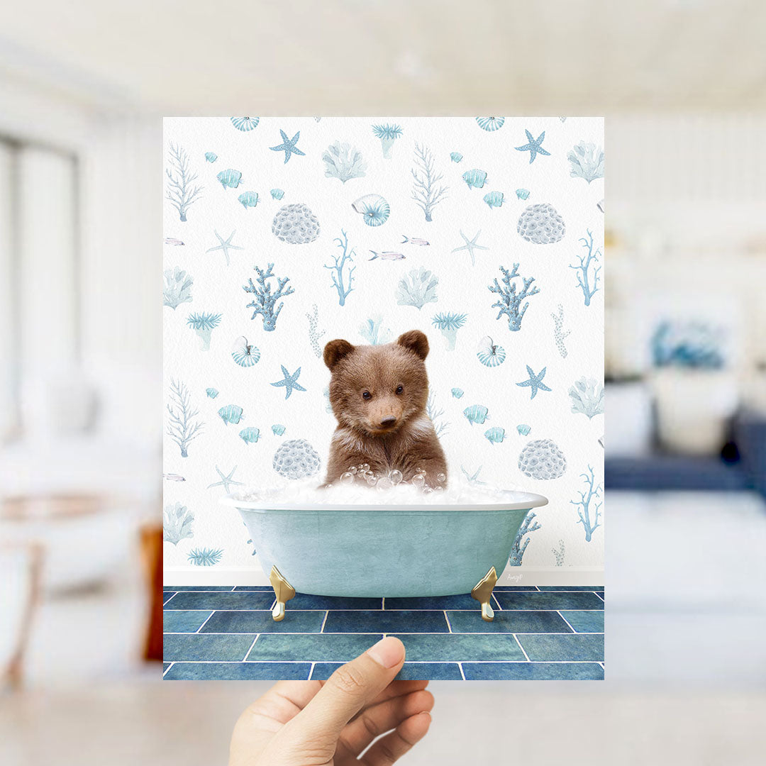 A hand is holding up a print featuring a small brown bear sitting in a blue bathtub filled with bubbles, surrounded by a pattern of seashells, starfish, and other marine life.