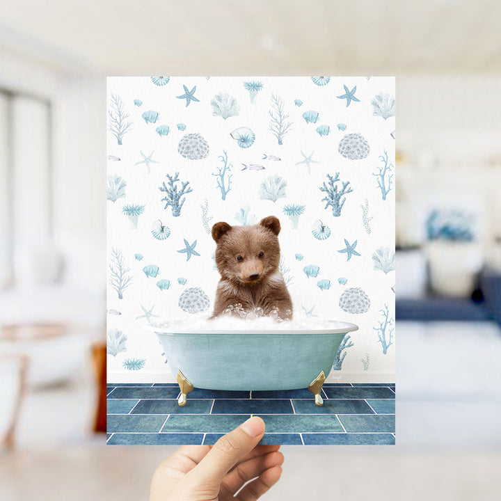 A hand is holding up a print featuring a small brown bear sitting in a blue bathtub filled with bubbles, surrounded by a pattern of seashells, starfish, and other marine life.