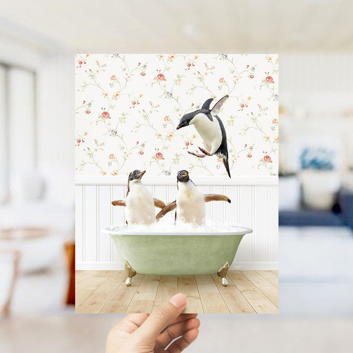 A hand holds up a framed photograph of two penguins sitting in a bathtub, with one penguin flying towards the other.