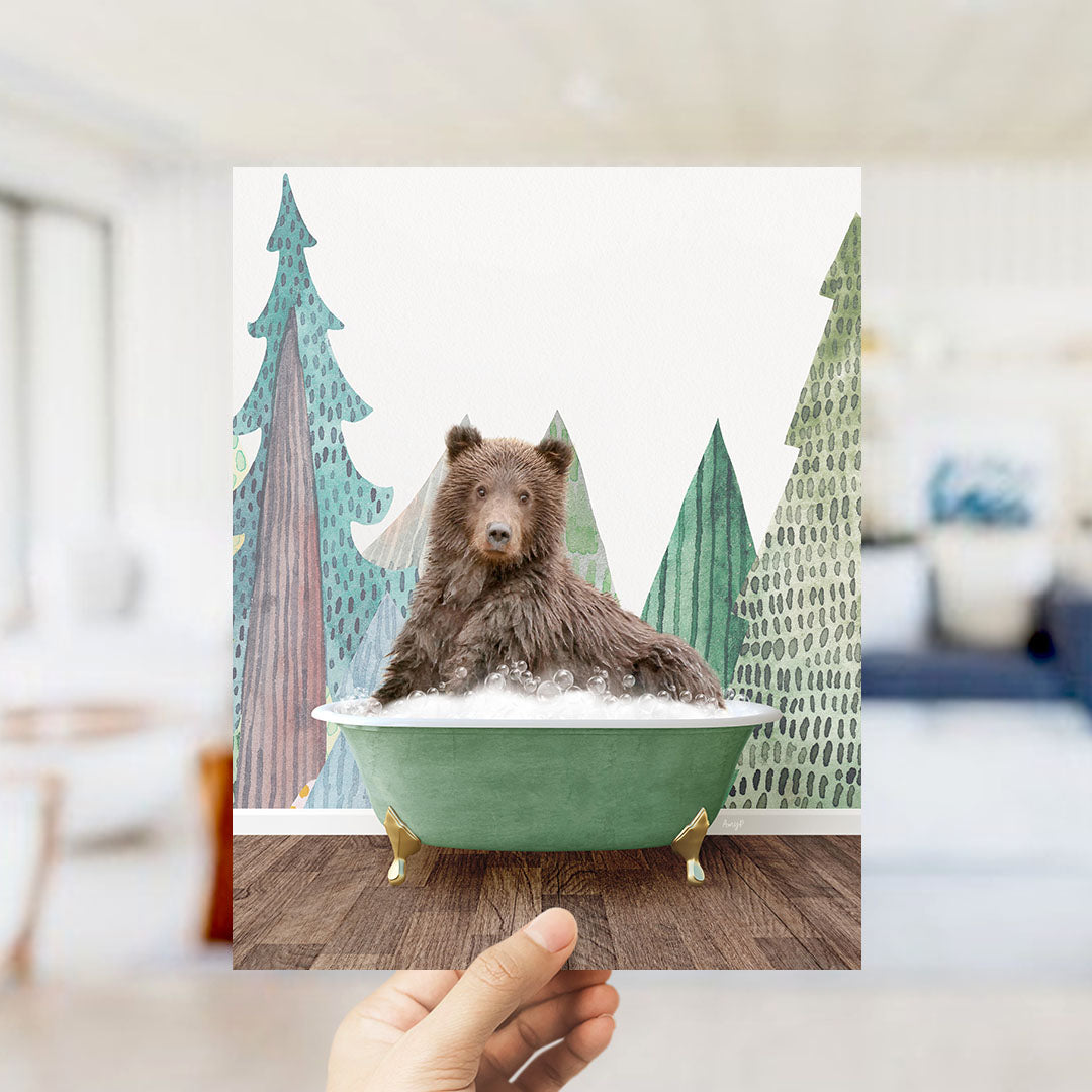 A hand holds up a framed image depicting a brown bear sitting in a green bathtub, surrounded by a forest scene with trees and a blue sky.
