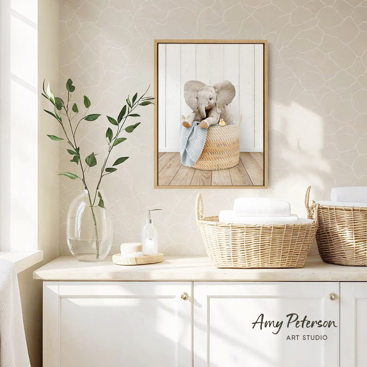 a framed picture of an elephant sitting in a basket, placed on a wall above a white cabinet with two woven baskets on top.