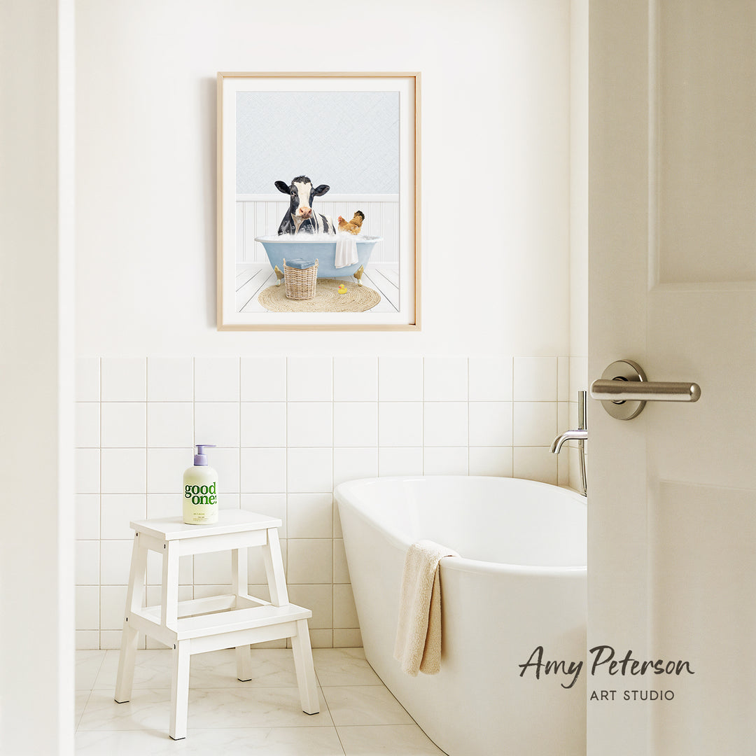 A framed image of a cow sitting in a bathtub is displayed on the wall above a white bathtub in a bathroom.