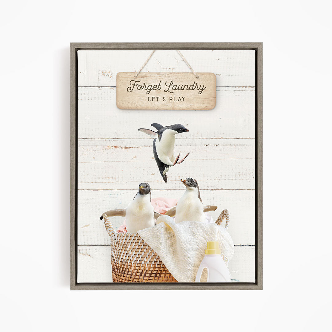A framed image depicting three penguins in a basket, with a sign that says "Forget Laundry, Let's Play" hanging above them.