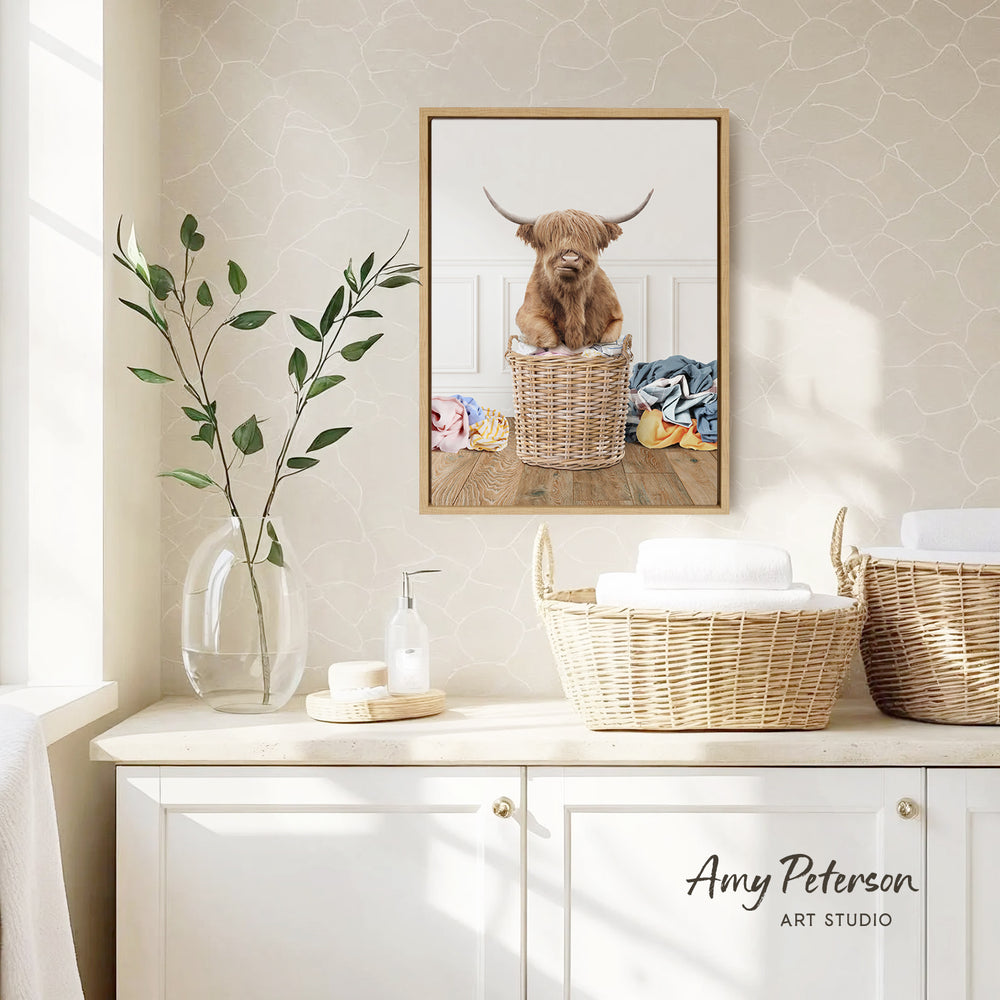 a framed photograph of a Highland cow sitting in a wicker basket, placed on a wall above a white cabinet.