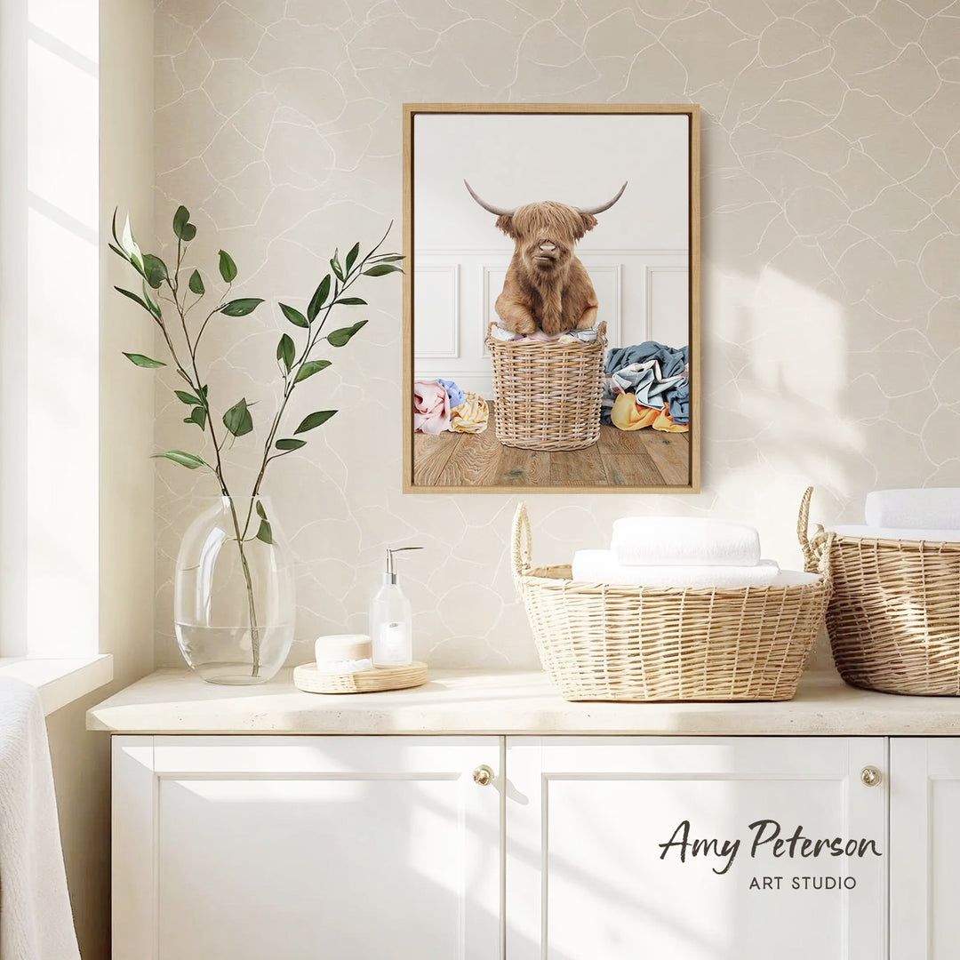 a framed photograph of a Highland cow sitting in a wicker basket, placed on a wall above a white cabinet.
