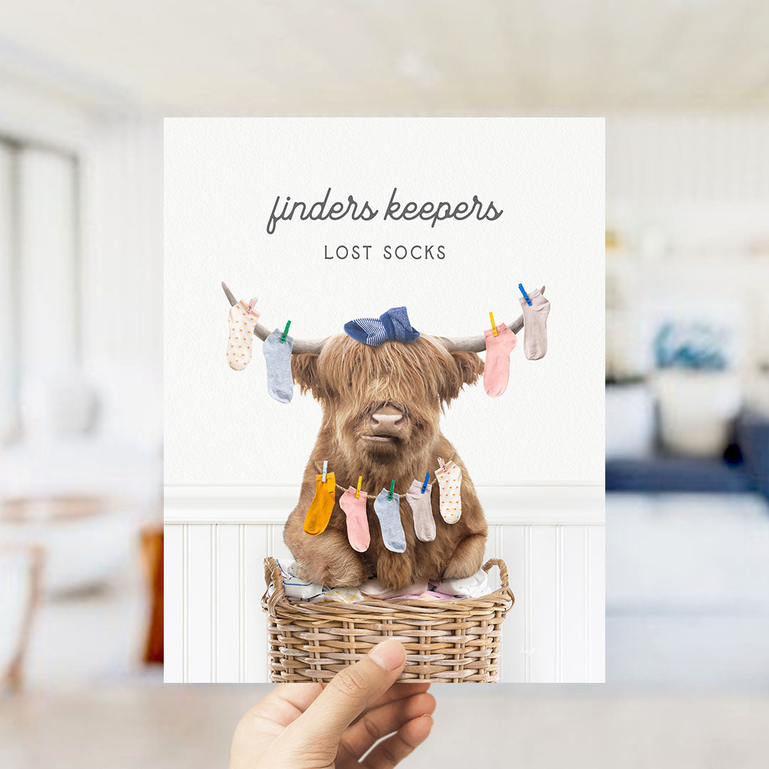 A hand holding a card with a cartoon image of a furry animal, likely a yak or a similar creature, sitting in a wicker basket filled with colorful socks.