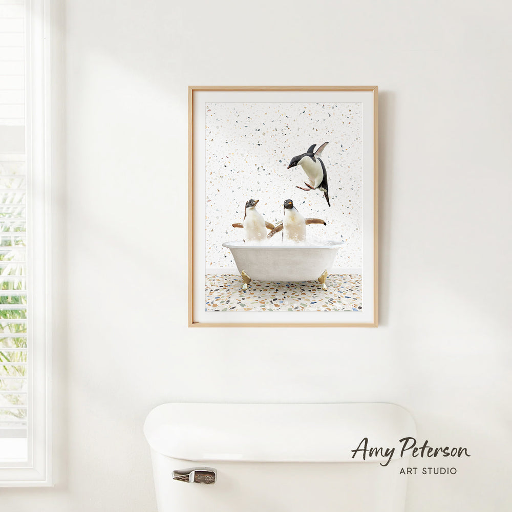 A framed image of two penguins sitting in a bathtub, surrounded by a speckled background, is hanging on a white wall.