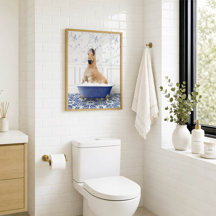 A bathroom with a white toilet, a framed picture of a dog sitting in a blue bathtub, and a window with greenery outside.