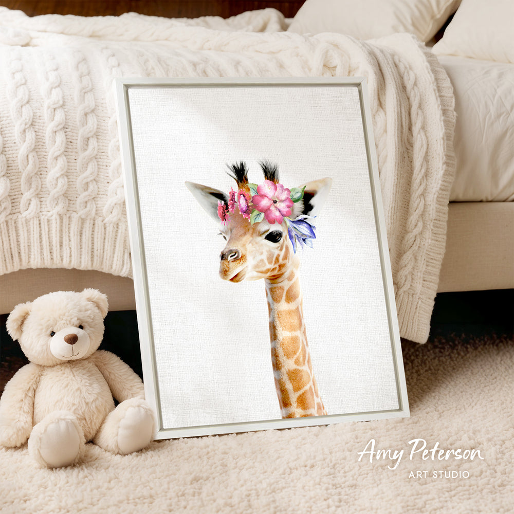 A framed print of a giraffe with a flower crown is displayed on a white bedspread, accompanied by a teddy bear on the floor.