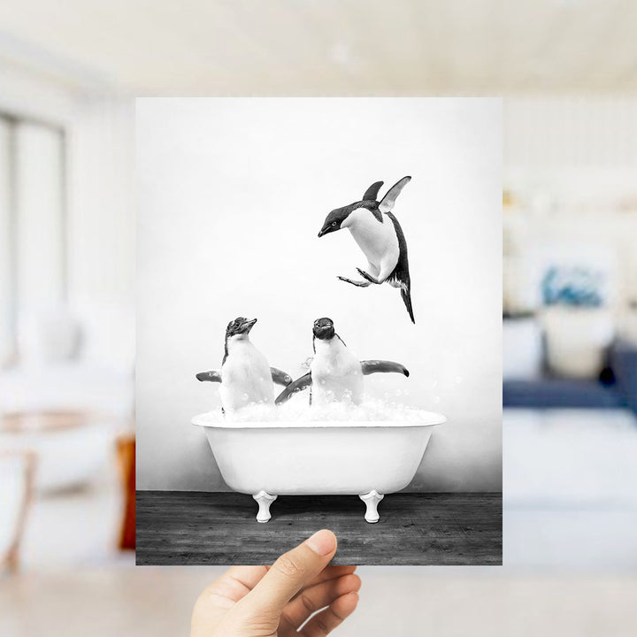 A hand holds up a framed photograph of two penguins sitting in a bathtub, with one penguin flying towards the other.