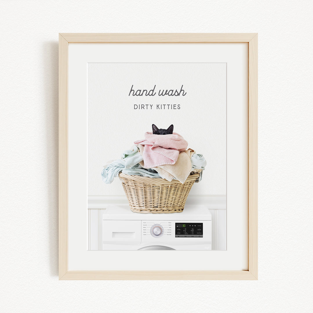 A framed image of a cat sitting in a basket filled with laundry, with the text "hand wash dirty kittens" written above the basket.