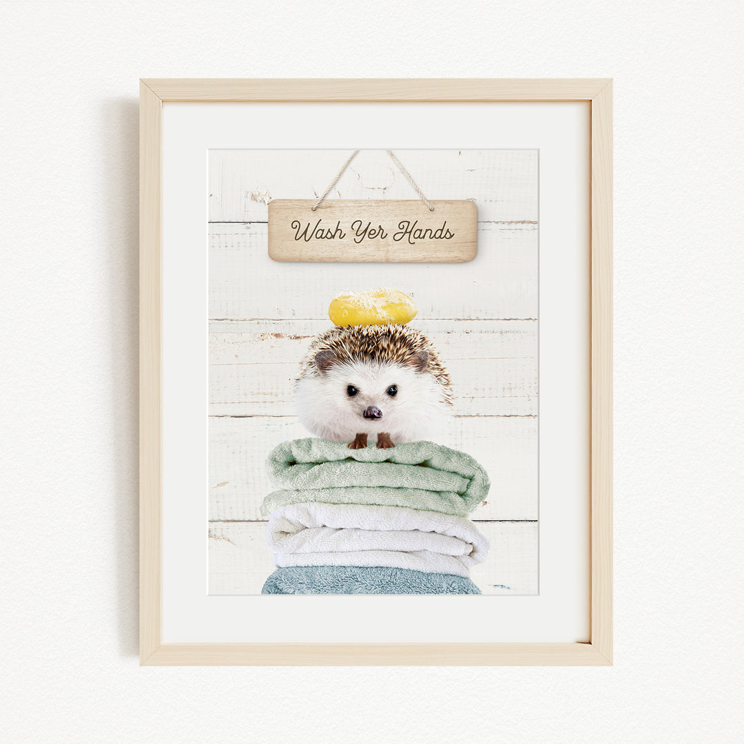 A framed image of a hedgehog sitting atop a stack of folded towels, with a wooden sign above that reads "Wash Your Hands".