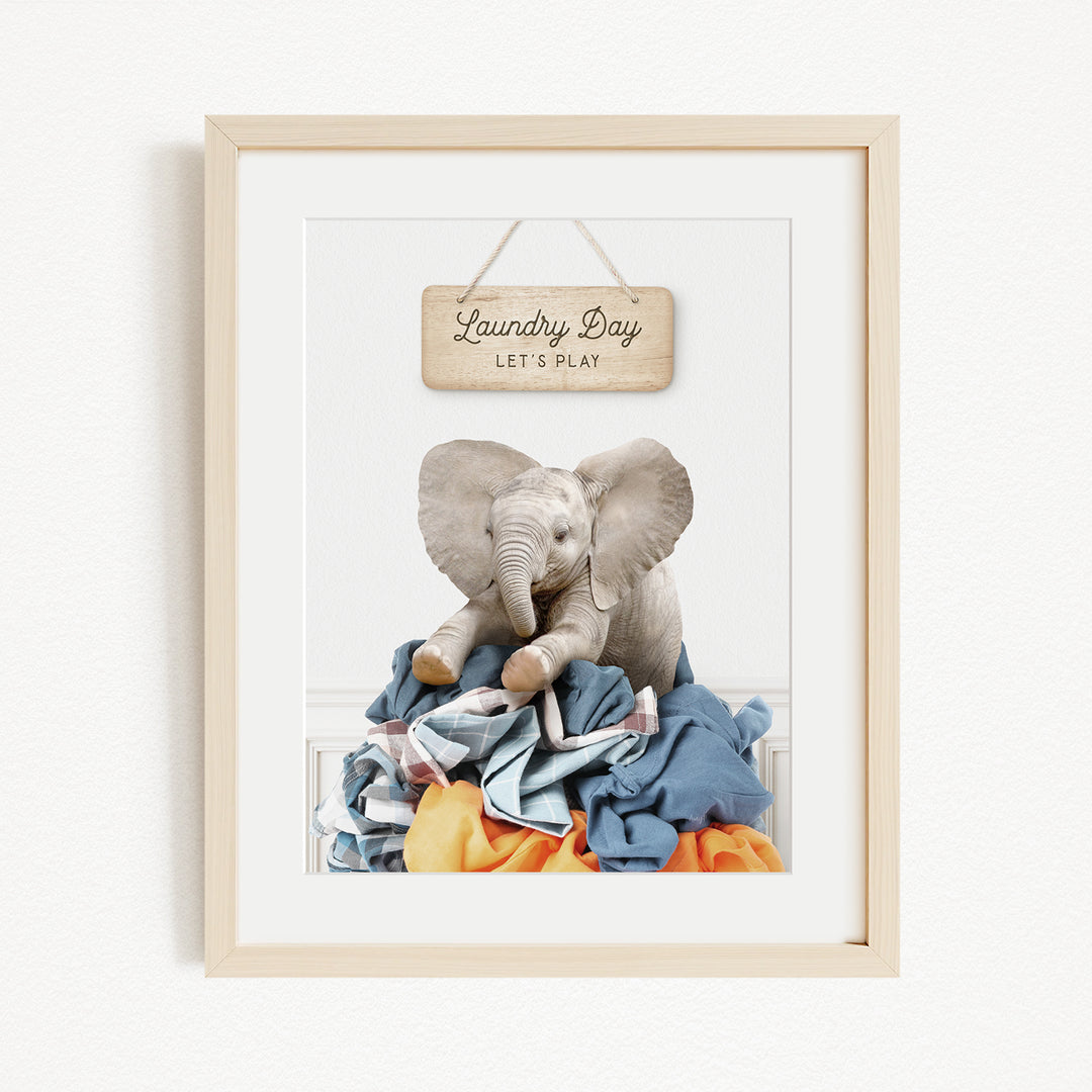 A framed image of a stuffed elephant sitting atop a pile of laundry, with a wooden sign above that reads "Laundry Day Let's Play".