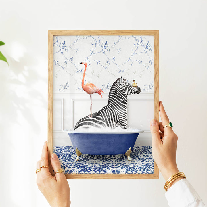 A person's hands are holding a framed picture of a zebra sitting in a blue bathtub, with a pink flamingo standing on the zebra's back.