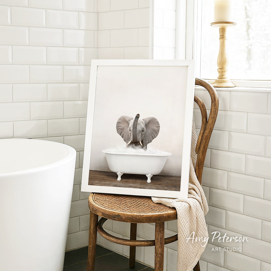 A framed photograph of an elephant sitting in a white bathtub is displayed on a wooden chair in a bathroom setting.