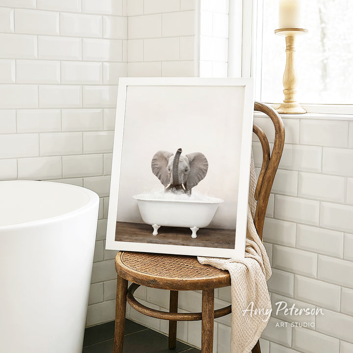 A framed photograph of an elephant sitting in a white bathtub is displayed on a wooden chair in a bathroom setting.