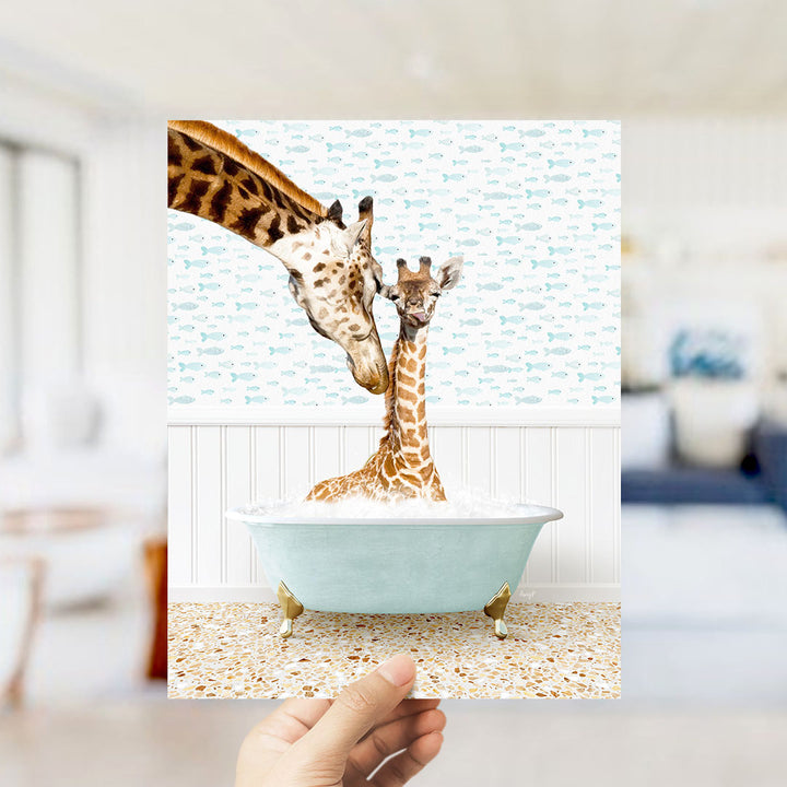 A hand holds up a framed image depicting two giraffes in a bathtub, with one giraffe partially submerged in the water and the other standing on the edge of the tub.