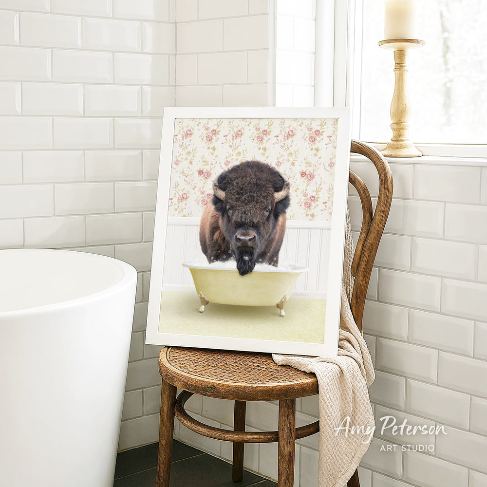 A framed image of a bison sitting in a bathtub is displayed on a wooden chair in a bathroom setting.