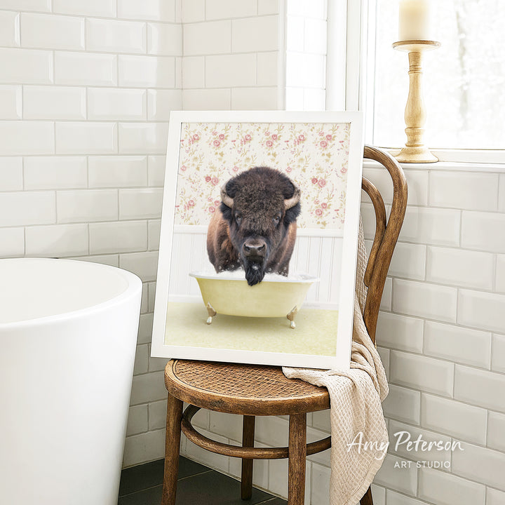 A framed image of a bison sitting in a bathtub is displayed on a wooden chair in a bathroom setting.