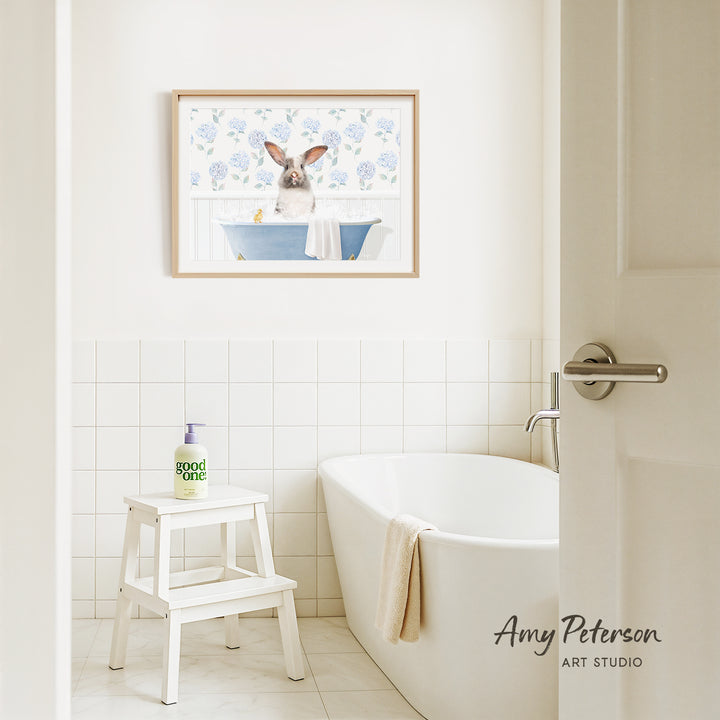 A framed painting of a rabbit sitting in a bathtub is hanging on a white tiled wall above a white bathtub in a bathroom.