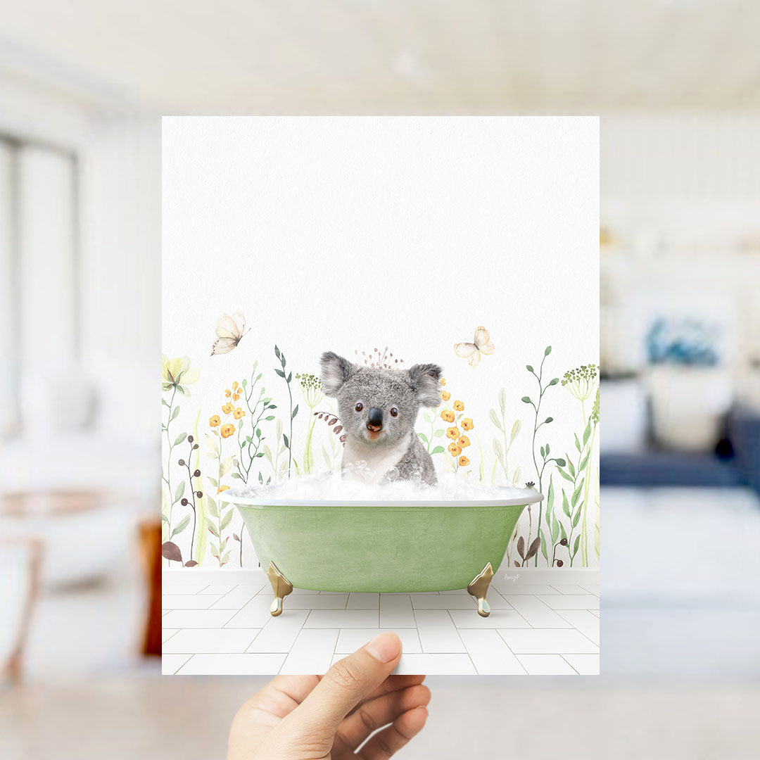 A hand holds up a framed image depicting a koala sitting in a green bathtub, surrounded by a floral and butterfly-themed background.