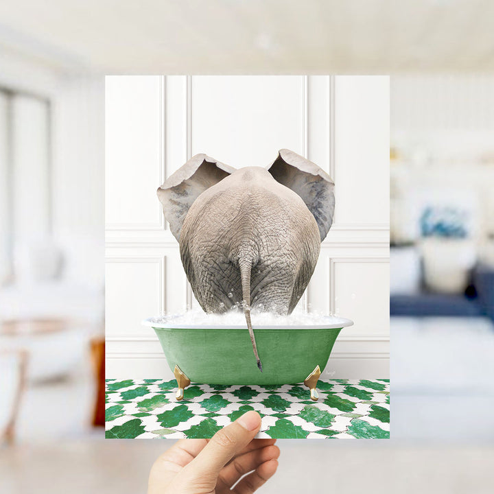 A hand holds up a framed image of an elephant sitting in a green bathtub filled with water, surrounded by a patterned floor.