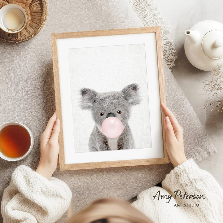 A person is holding a framed picture of a koala bear blowing a bubble with a pink bubblegum bubble.