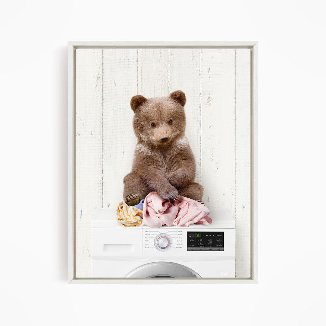 A brown bear sitting on top of a white washing machine, with clothes and other laundry items around it.