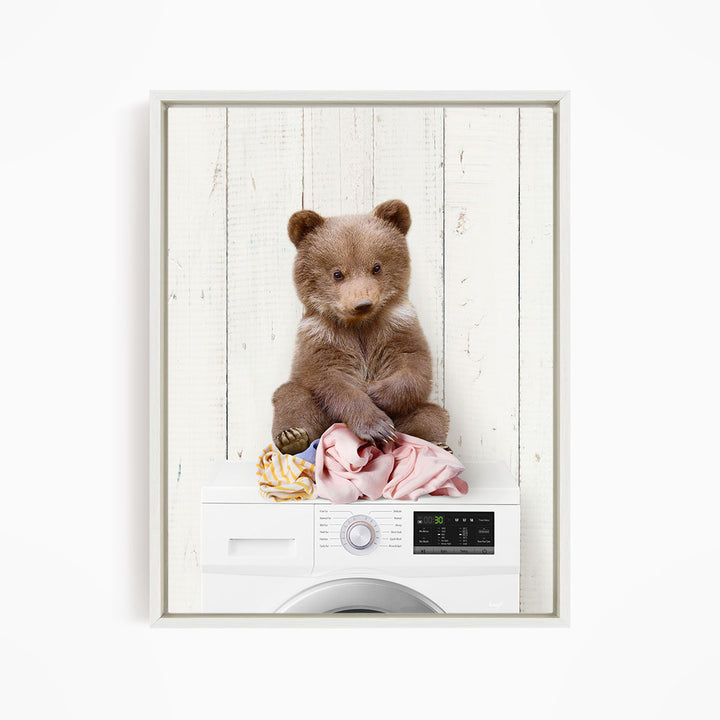 A brown bear sitting on top of a white washing machine, with clothes and other laundry items around it.