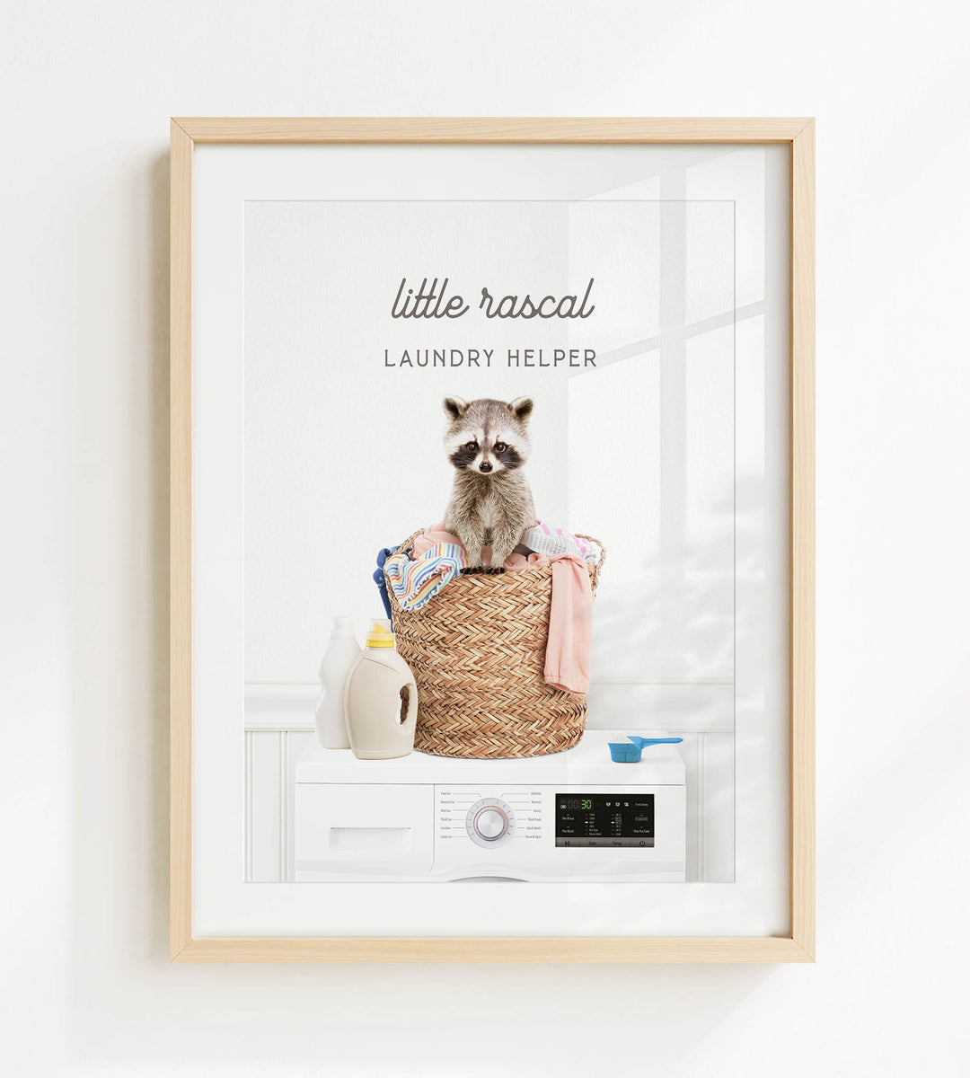 A framed image depicting a raccoon sitting on a basket filled with laundry, including a bottle of detergent and a measuring cup.