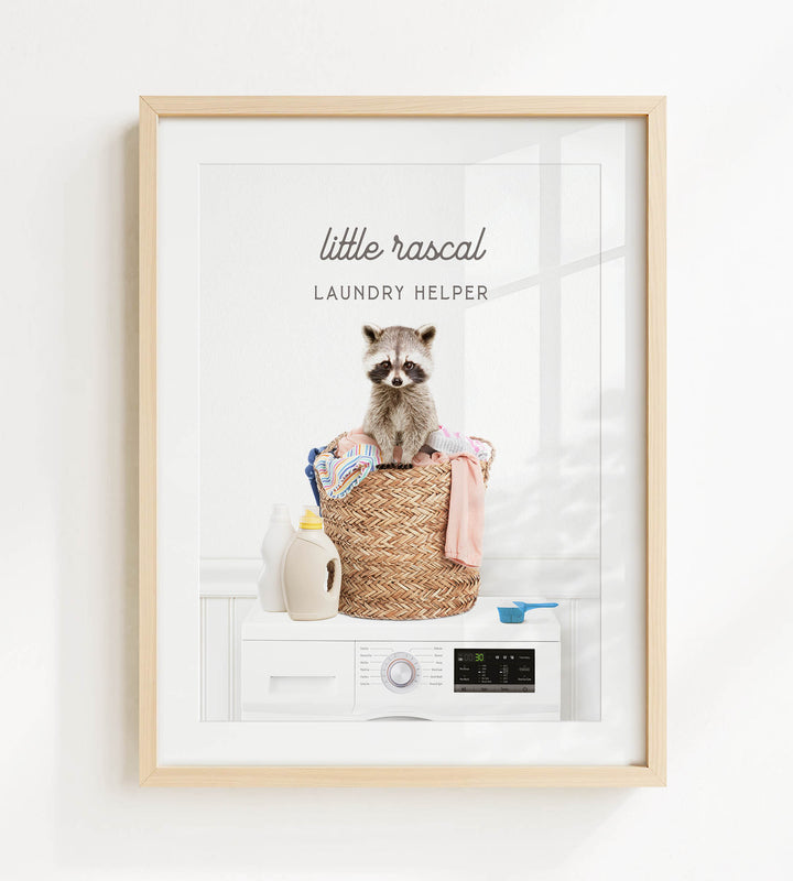 A framed image depicting a raccoon sitting on a basket filled with laundry, including a bottle of detergent and a measuring cup.