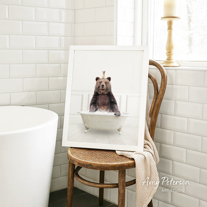 A framed image of a bear sitting in a bathtub, with a crown on its head, is displayed on a wooden chair in a bathroom setting.