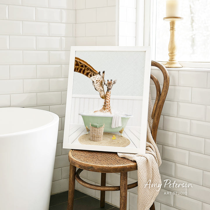 A framed image of two giraffes sitting in a bathtub, with one giraffe appearing to be bathing the other giraffe. The image is displayed on a wooden chair in a bathroom setting.