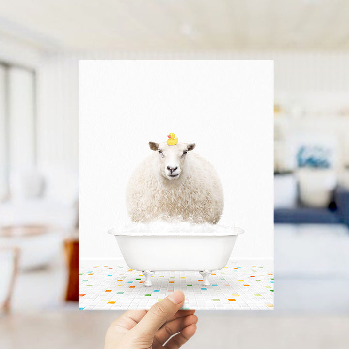 A hand holds up a framed image depicting a fluffy white sheep sitting in a white bathtub filled with bubbles, with a yellow rubber duck perched on its head.