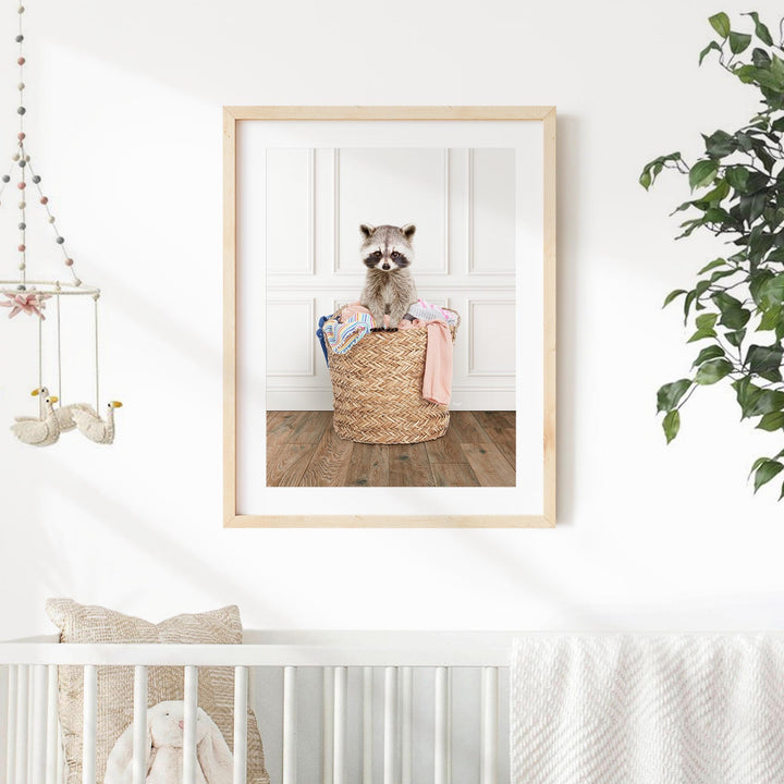 A framed photograph of a raccoon sitting in a woven basket on a wooden floor, surrounded by various baby items.