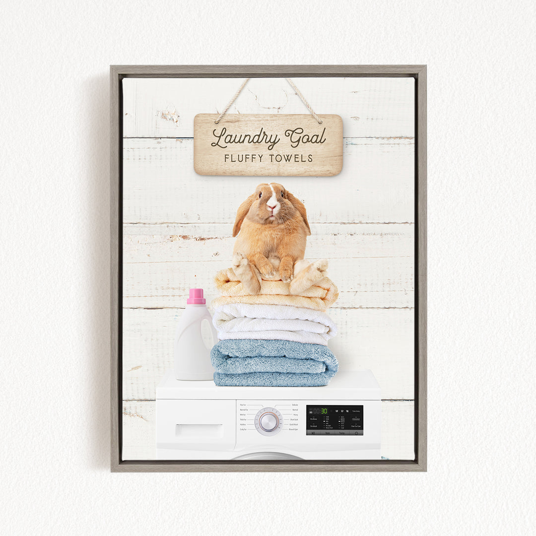 A framed image of a rabbit sitting atop a stack of fluffy towels, with a sign above that reads "Laundry Goal Fluffy Towels".