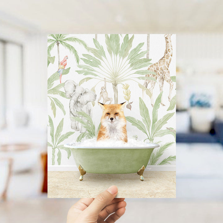 A hand holds up a framed print featuring a fox sitting in a bathtub surrounded by tropical plants and animals, including a giraffe and a parrot.