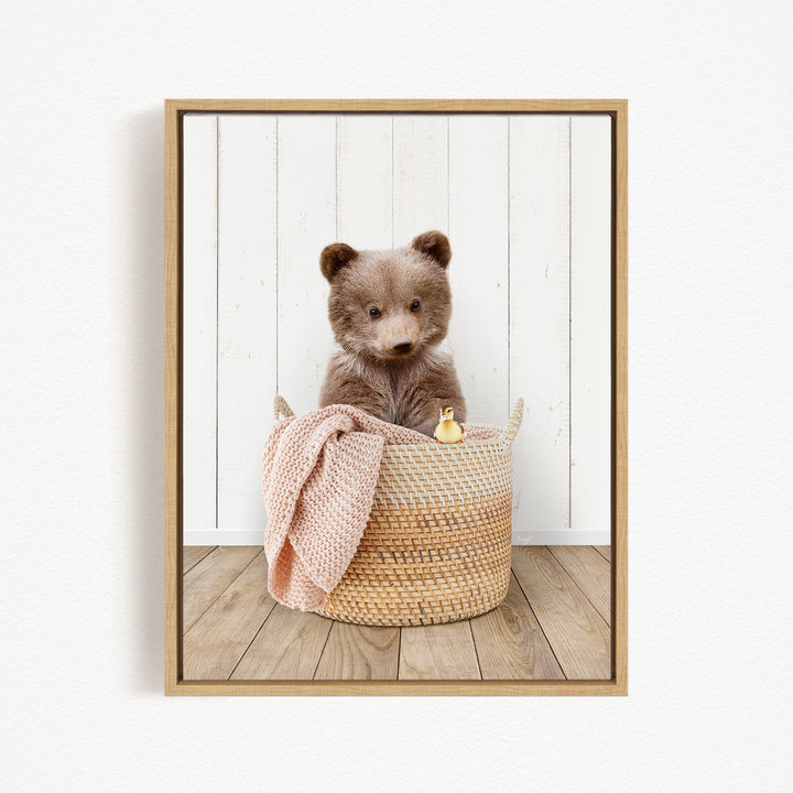 A brown bear sitting in a woven basket, with a pink blanket and a small yellow object nearby.