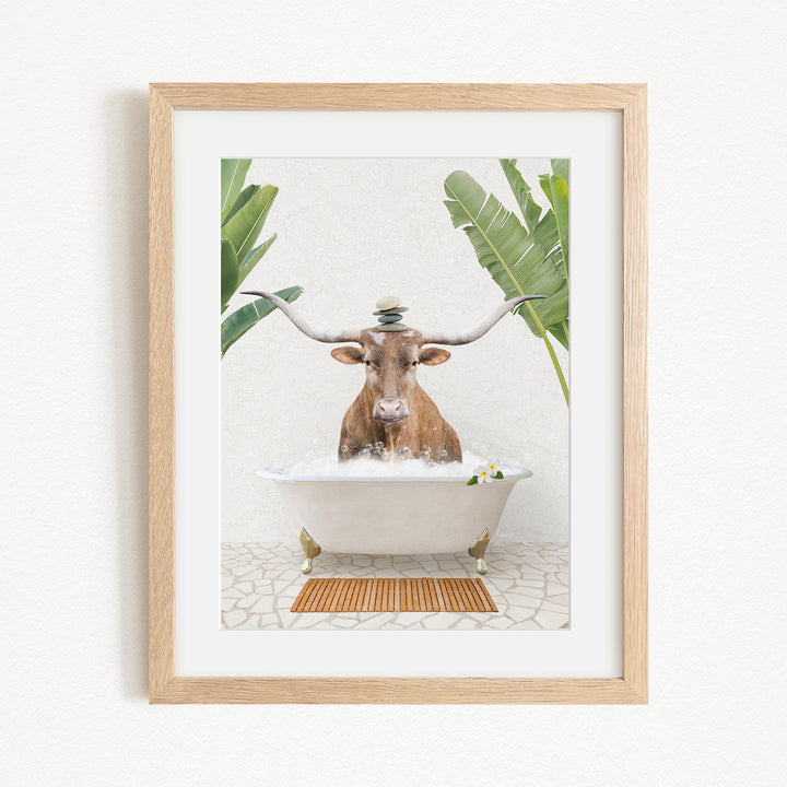 A framed image depicting a cow sitting in a bathtub filled with water, surrounded by tropical plants.