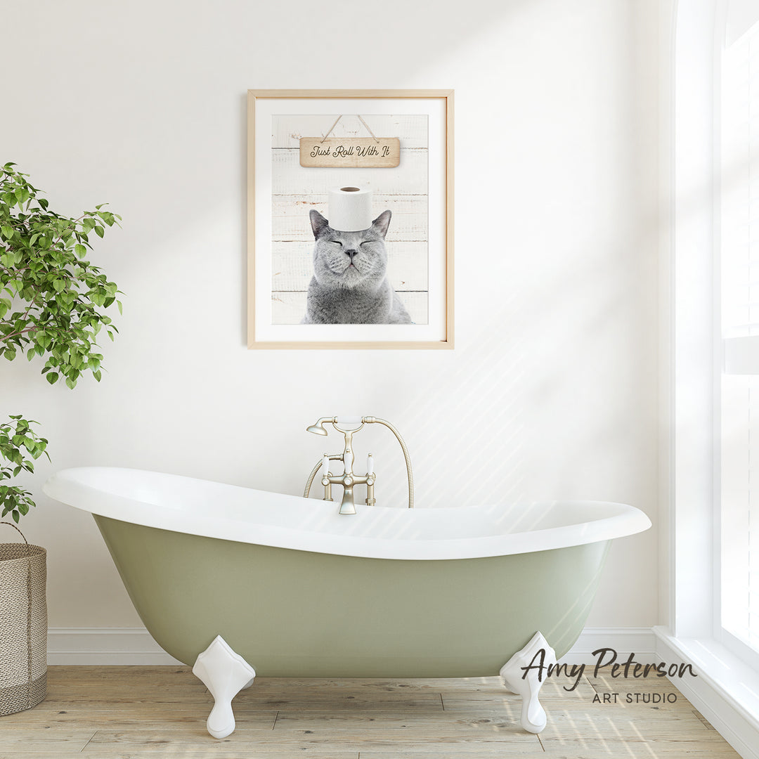A framed picture of a cat with a humorous caption hangs on a white wall above a green clawfoot bathtub in a bright, airy bathroom.