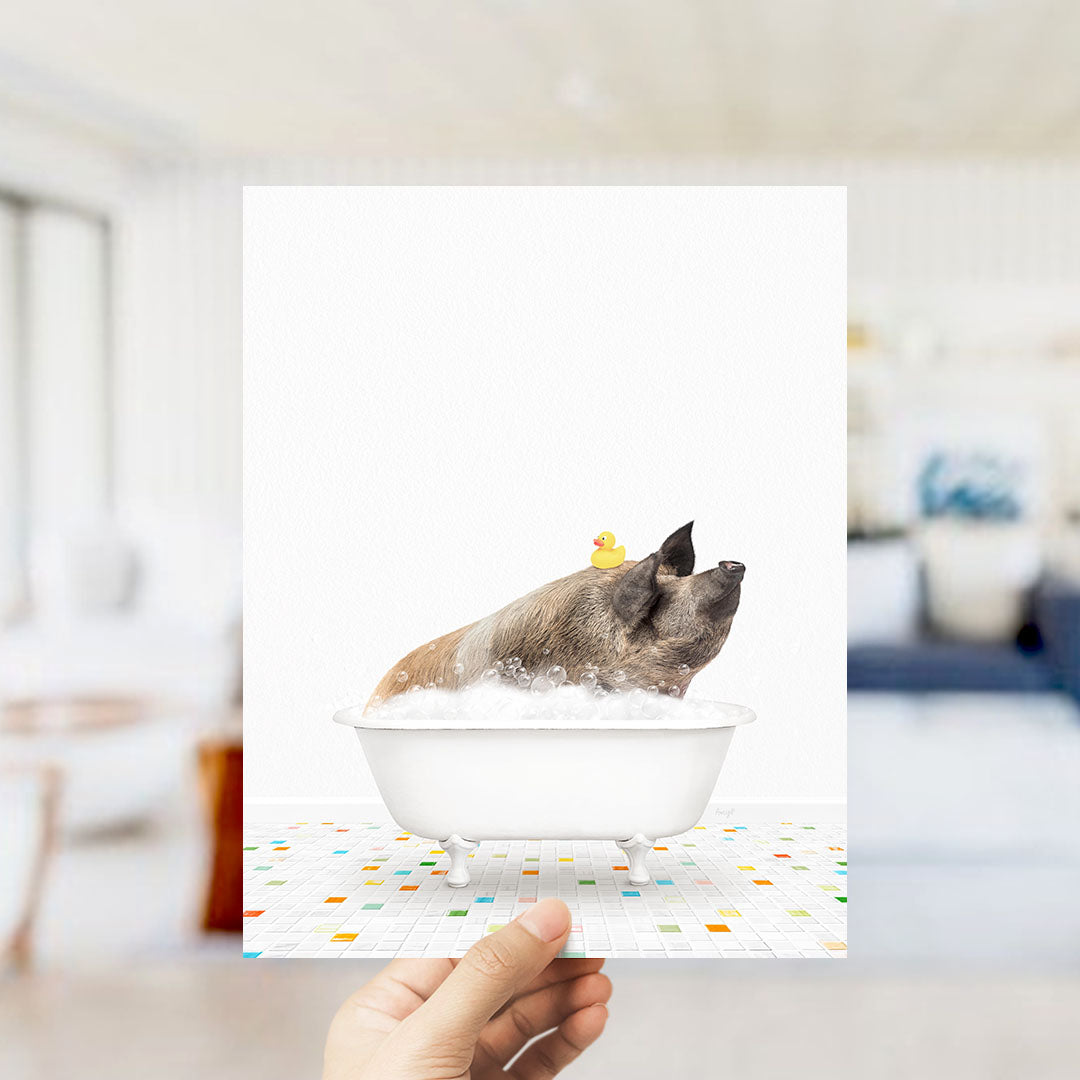 A hand holds up a framed image depicting a pig sitting in a bathtub, with a rubber duck on its head.
