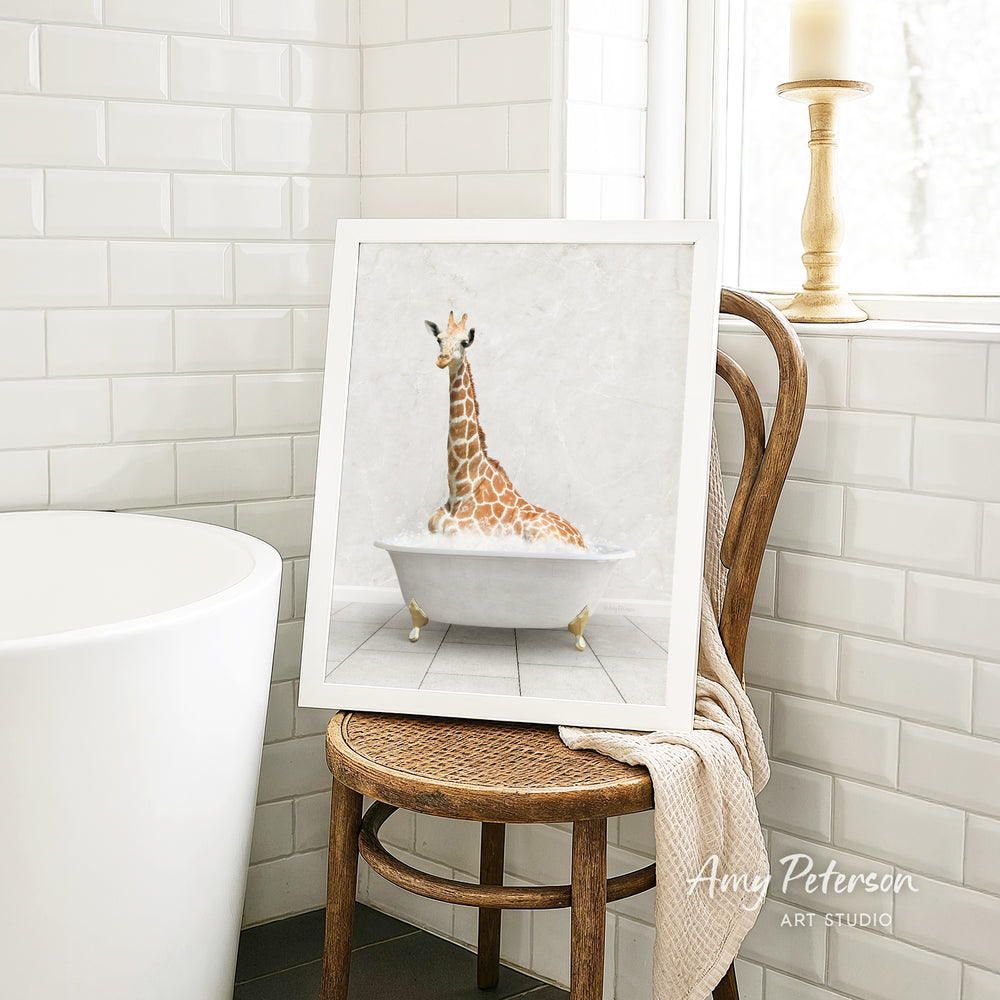 A framed image of a giraffe sitting in a bathtub is displayed on a wooden chair in a bathroom setting.