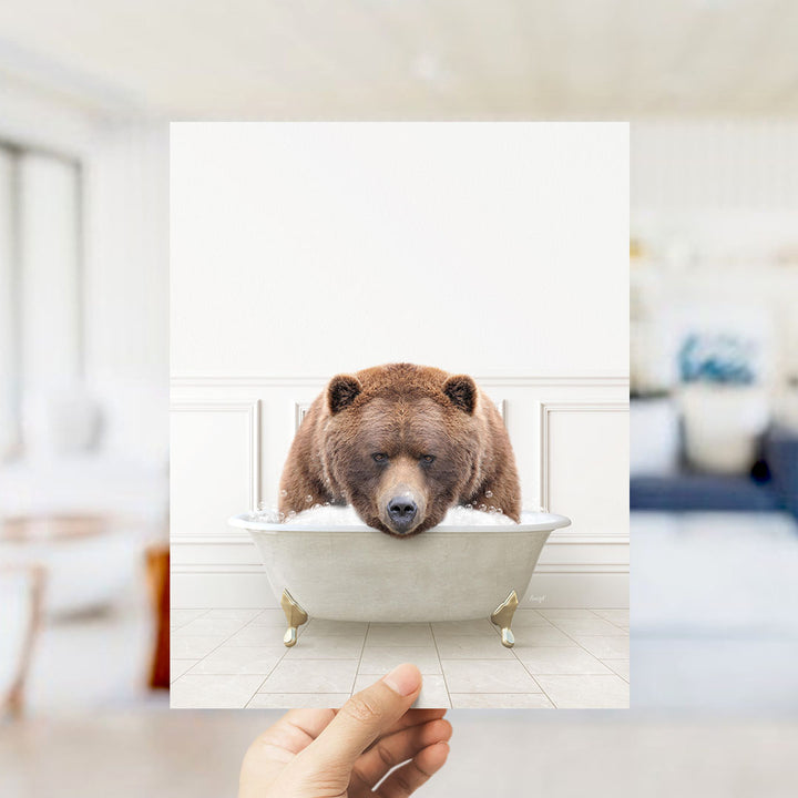 A hand holds up a framed image of a brown bear sitting in a bathtub filled with water, creating a whimsical and playful scene.