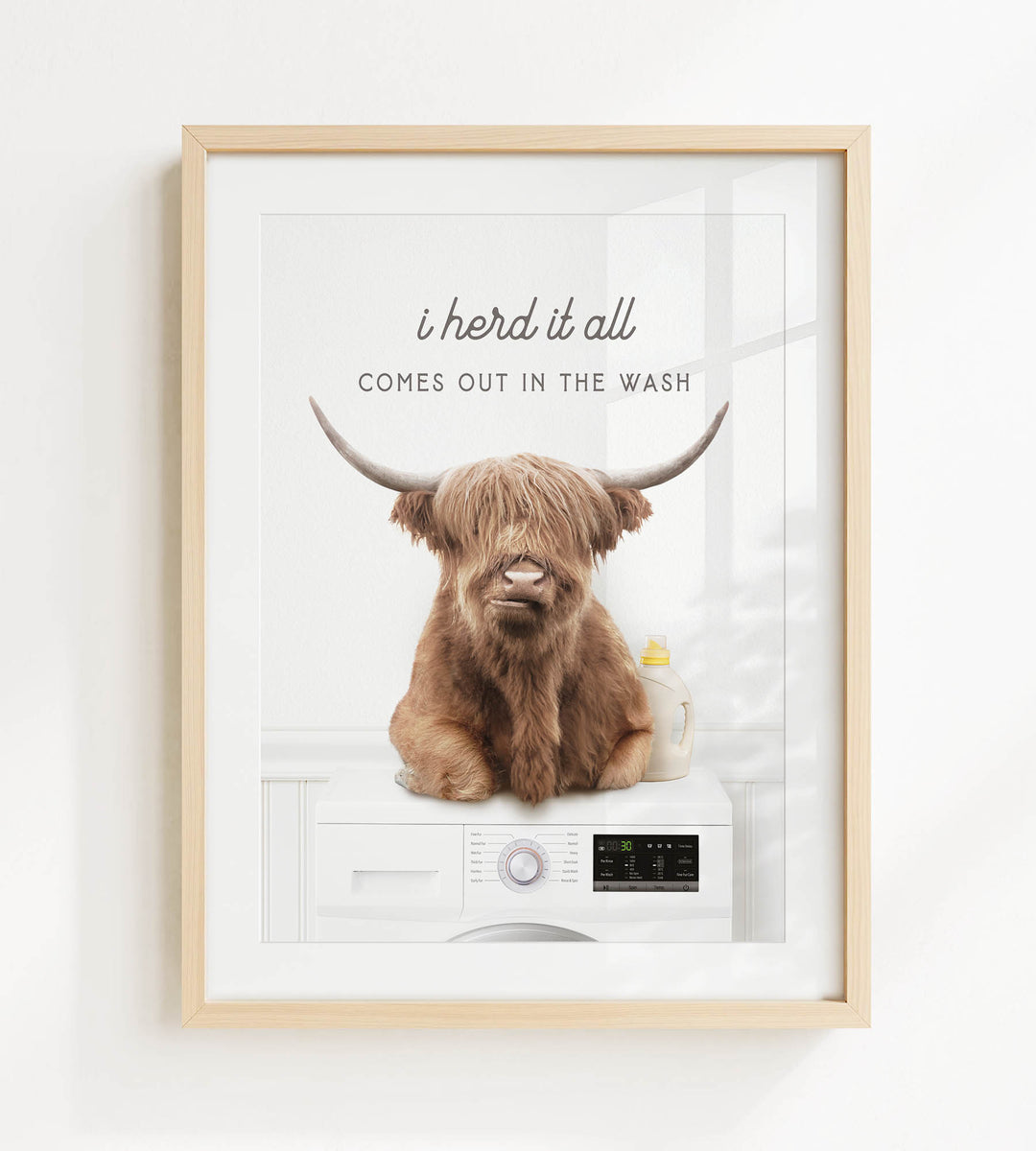 A framed image depicting a brown cow with horns sitting on a white washing machine, with the text "I herd it all comes out in the wash" above the cow.