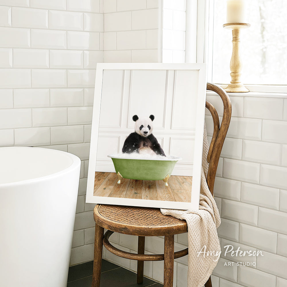 A framed image of a panda sitting in a green bathtub, with a towel draped over the back of a wooden chair in a bathroom setting.
