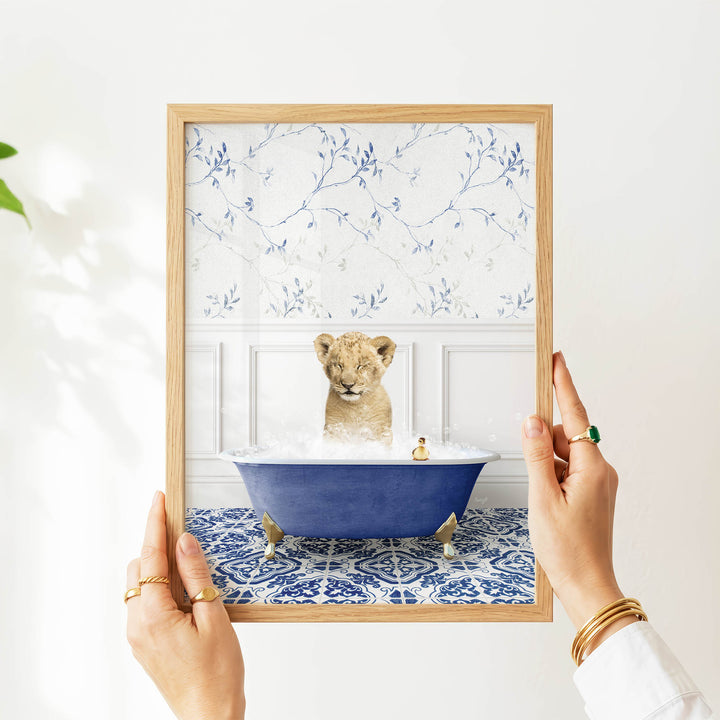 A person's hands are holding a framed picture of a lion sitting in a blue bathtub filled with water and bubbles, with a small rubber duck floating in the water.